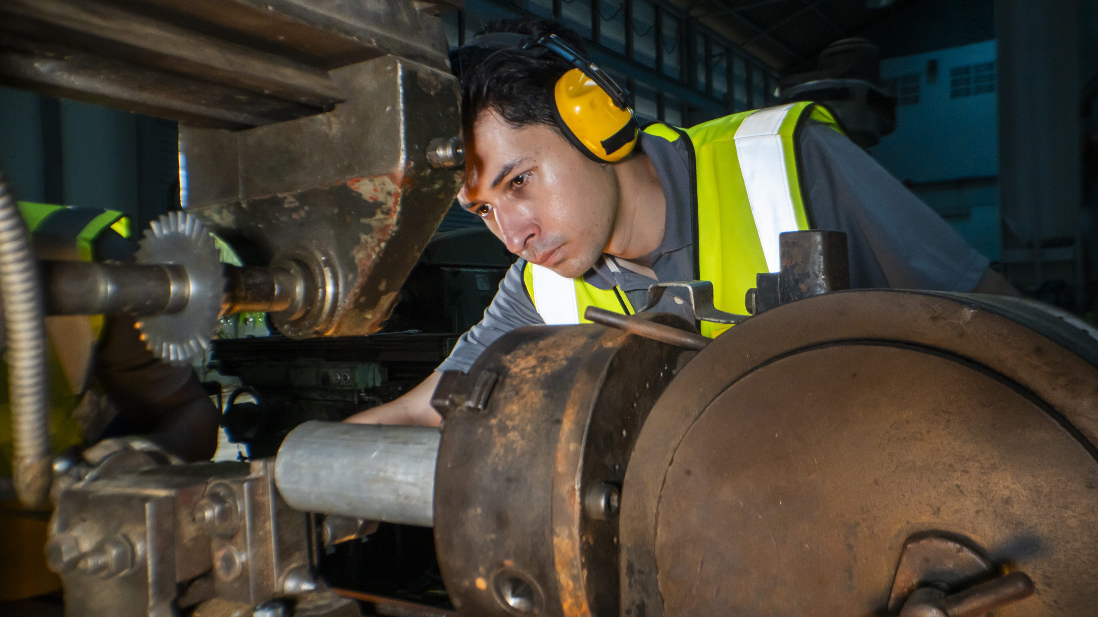 industrial engineer intently operates a gear driven machine in a workshop. Wearing ear protection and a reflective vest, he focuses on precision work, ensuring safe operation of the heavy equipment.